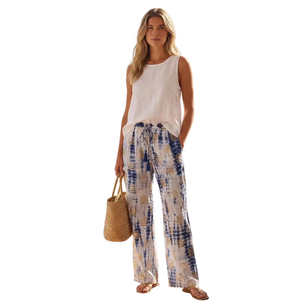 A woman stands against a solid white background wearing a sleeveless white top and wide-leg pants with a blue, white, and gold tie-dye pattern. She holds a woven straw tote bag and wears flat sandals.