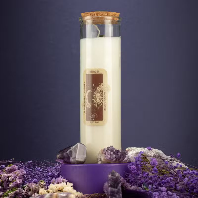 A tall glass jar candle with a cork stopper and cream-colored wax stands on a purple pedestal surrounded by purple flowers, crystals, and dried botanicals against a dark blue background.