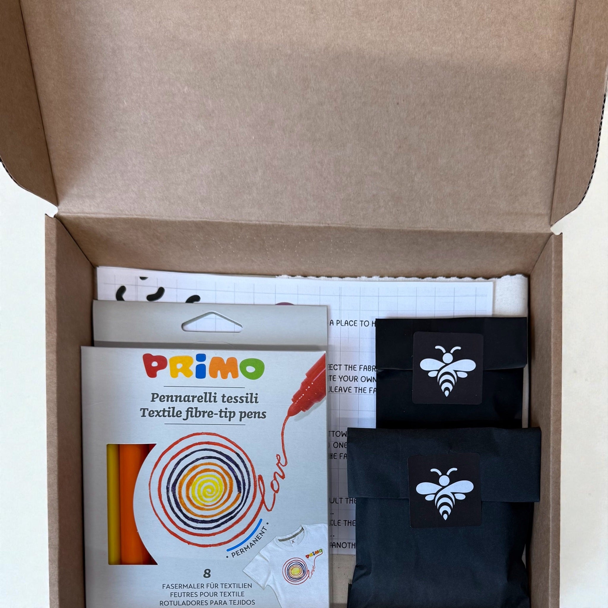 Open cardboard box containing Primo textile fibre-tip pens and black pouches with bee logo.