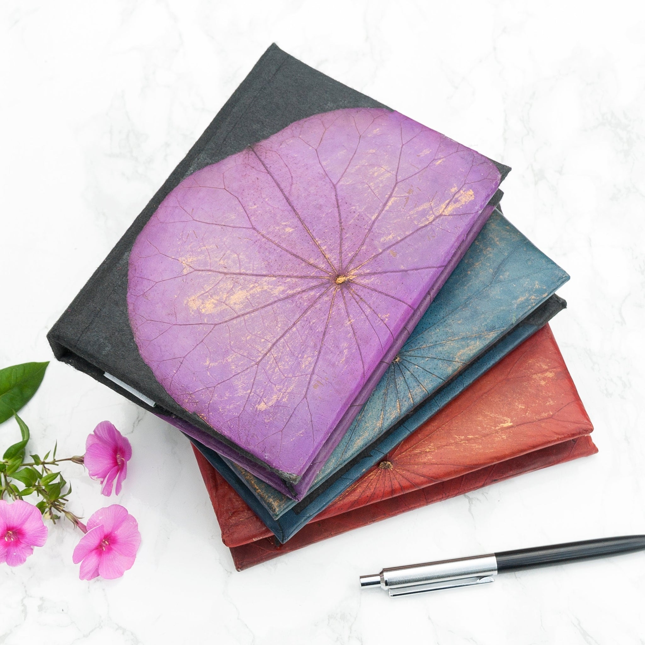 Stack of notebooks with floral designs on a white surface with flowers and a pen.