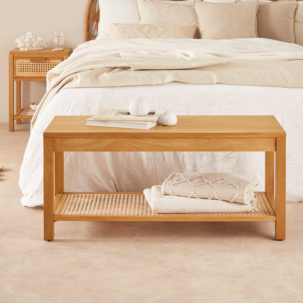 Wooden bench with a woven shelf in front of a bed with white bedding.