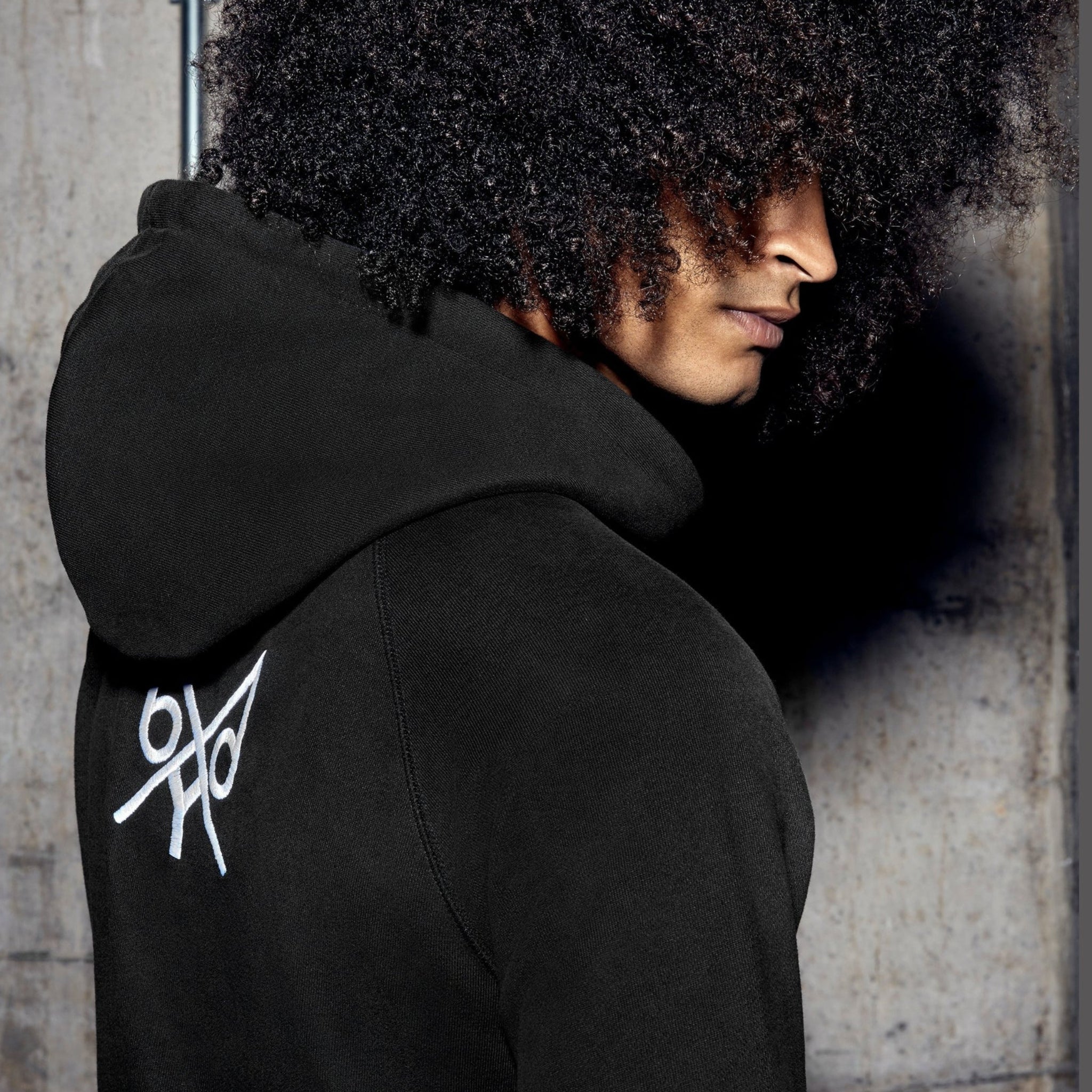 Person wearing a black hoodie with a white logo on the back, standing against a textured wall.