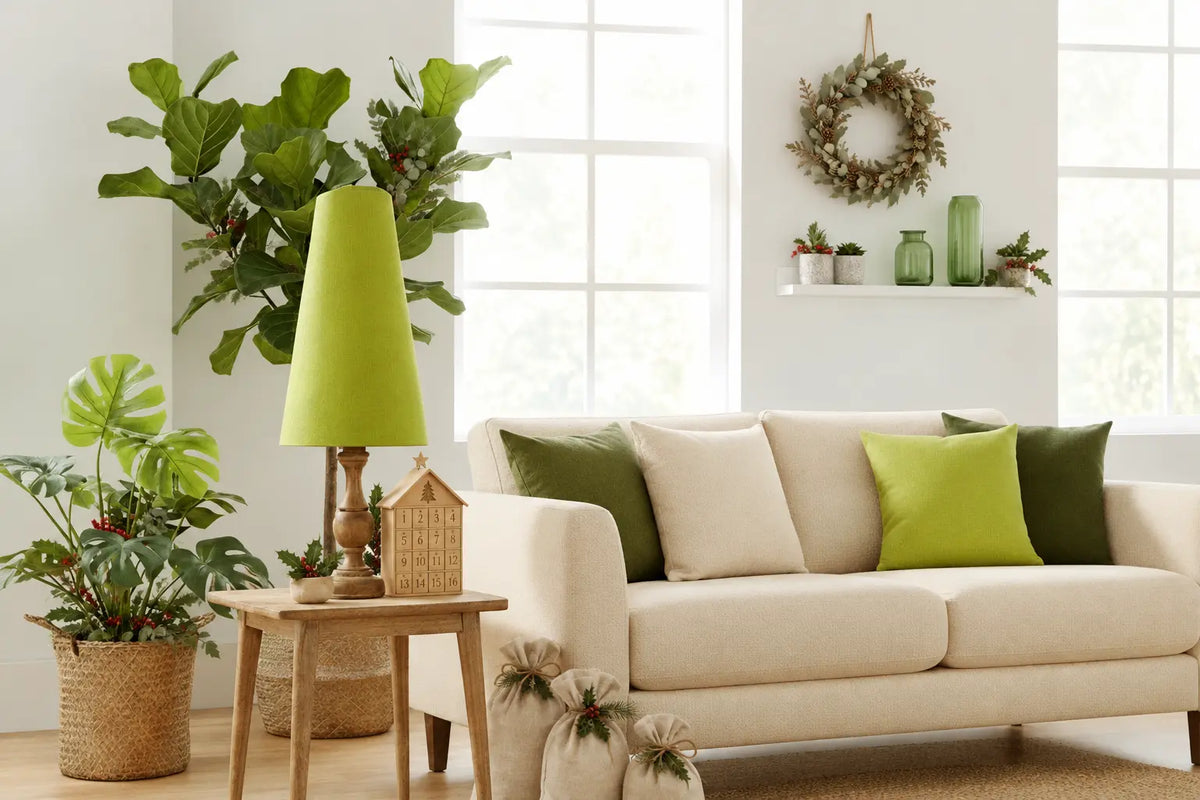A styled living room scene with eco-friendly Christmas decor, featuring a cream-colored sofa with green and olive accent pillows, a matching green table lamp on a wooden side table, and various potted plants against a bright white wall.