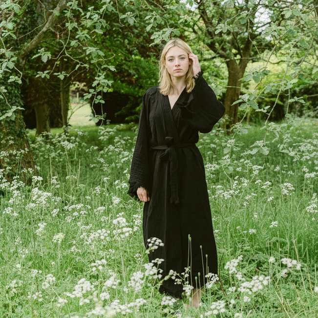 A woman wearing a long black organic cotton robe stands in a grassy field with white flowers and leafy trees in the background.