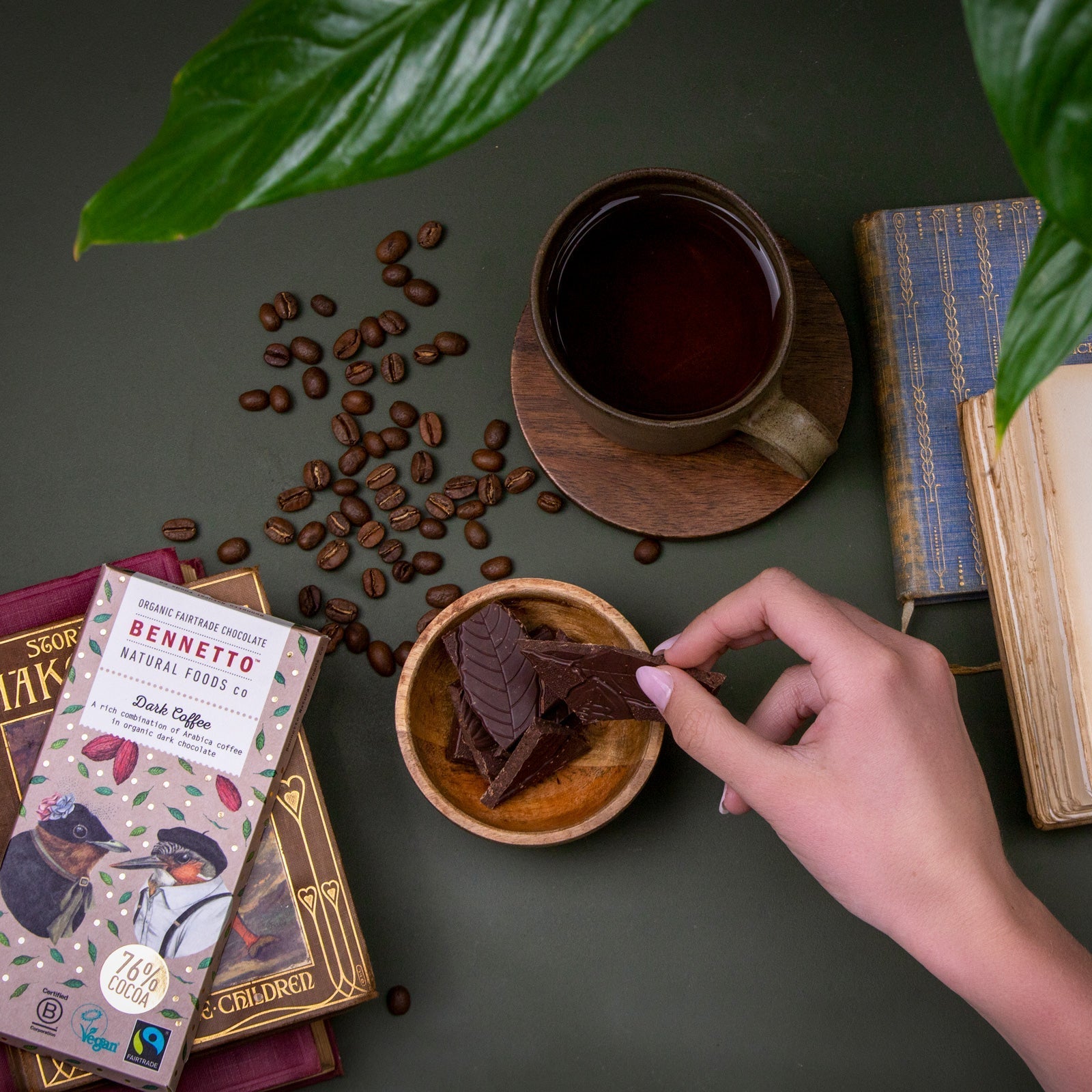 Hand holding a chocolate bar over a bowl of chocolate pieces with coffee beans, a cup of coffee, and books on a dark surface.