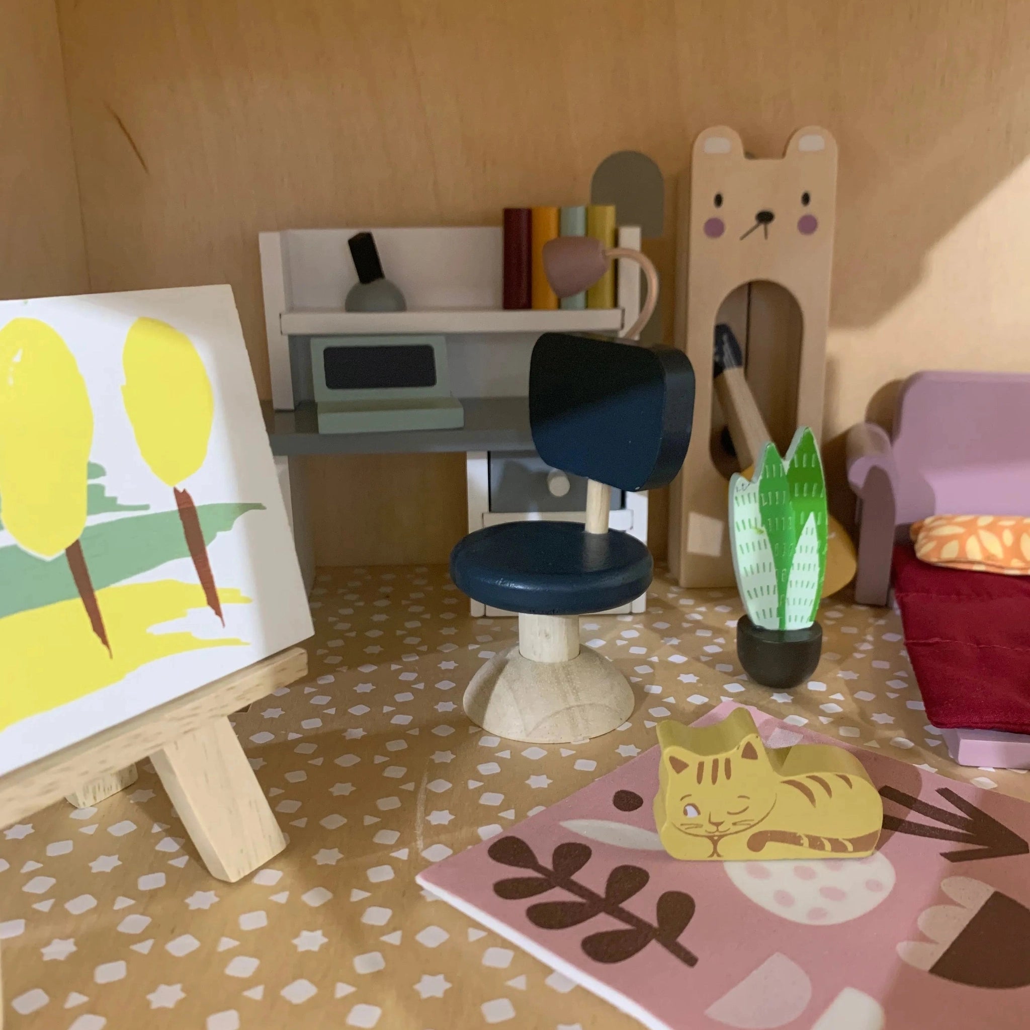 A doll's house study with toy furniture, books, and a painting on an easel.