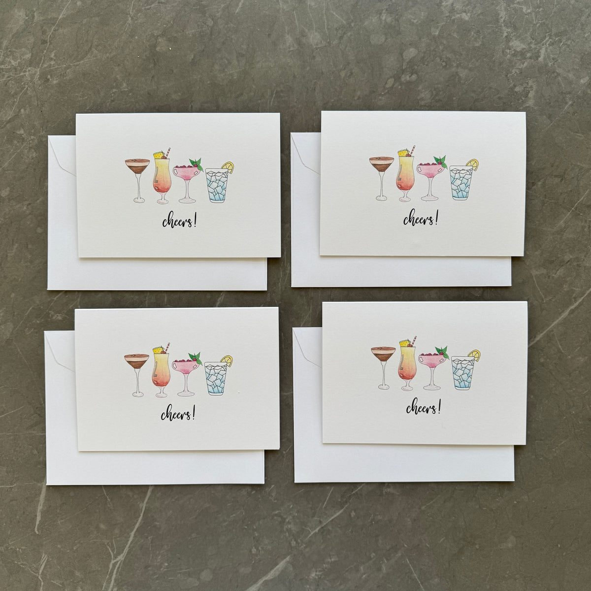 Set of four greeting cards with cocktail illustrations and 'cheers!' text on a gray surface.