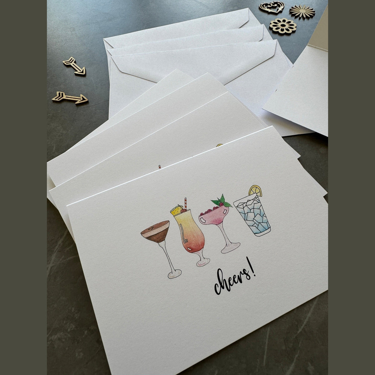 Set of greeting cards with cocktail illustrations and 'cheers!' text on a dark surface.