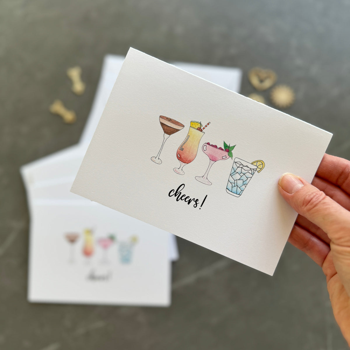 Hand holding a card with cocktail illustrations and 'cheers!' text on a gray surface.
