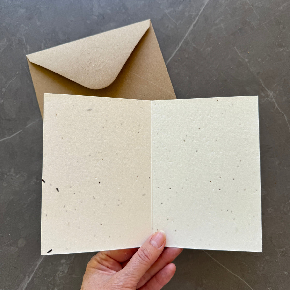 Open greeting card with textured pages held by a hand, next to a closed brown envelope on a dark surface.