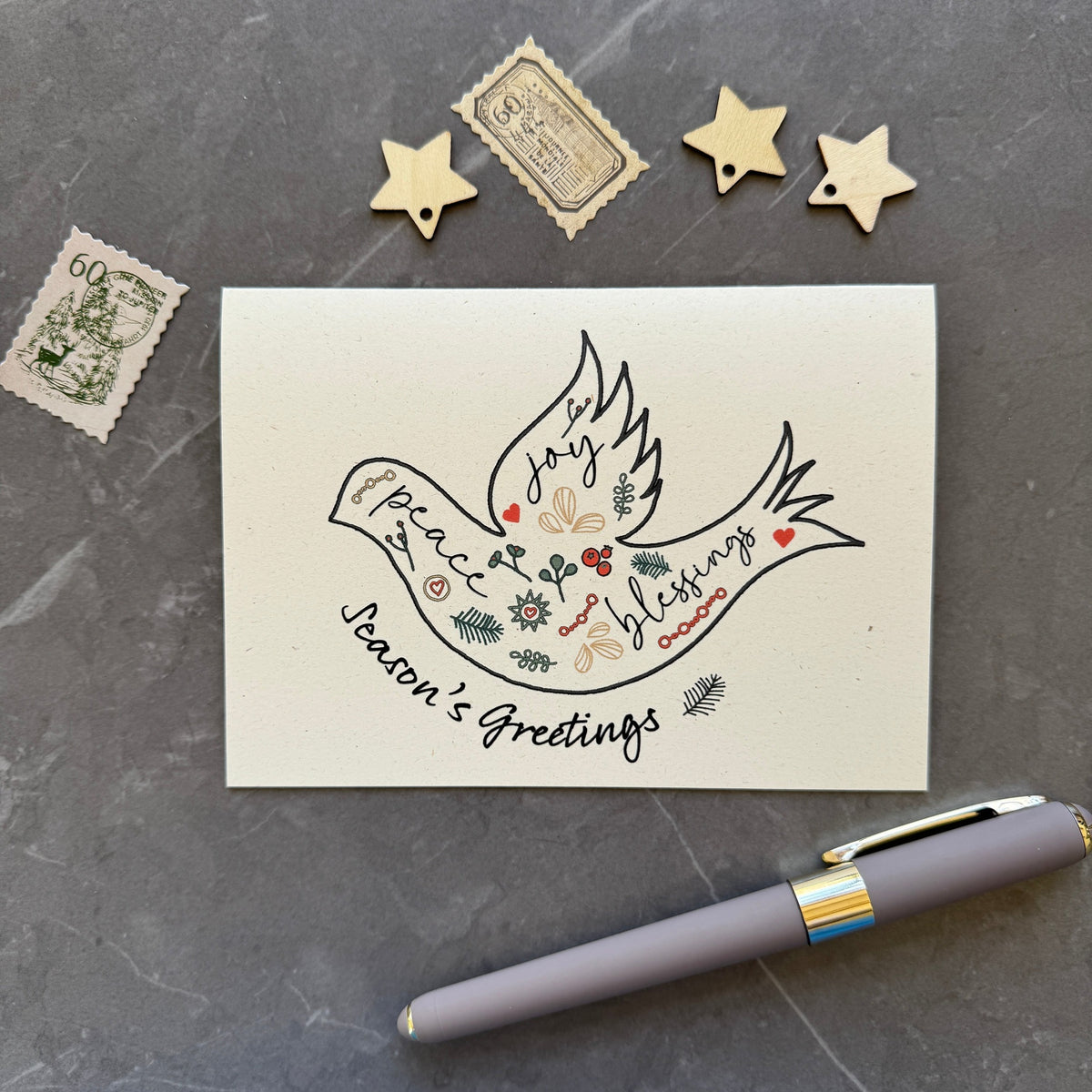 Season's Greetings card with decorative dove design on a gray surface with a pen and decorative items.