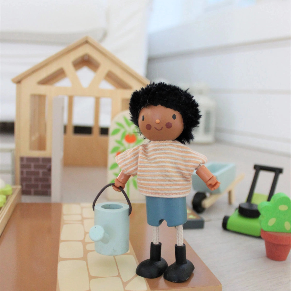 Doll holding a miniature blue watering can beside the Tender Leaf wooden greenhouse