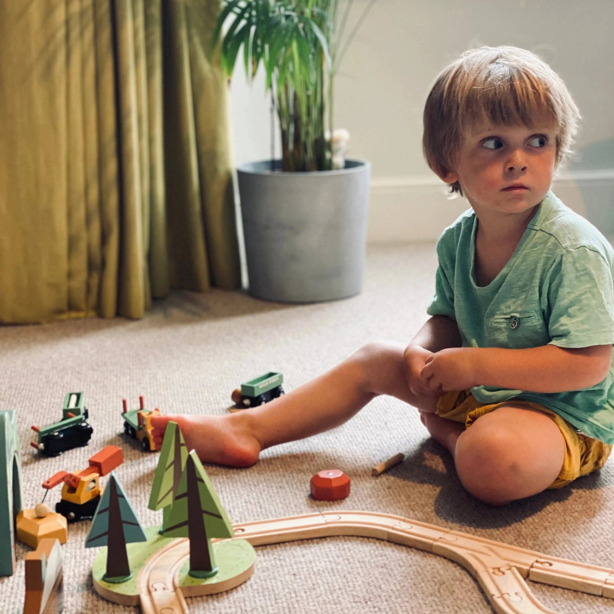 A young child sitting on a carpeted floor in a playroom, surrounded by wooden toy trains, trees, and a wooden train track. The child is wearing a green t-shirt and yellow shorts.