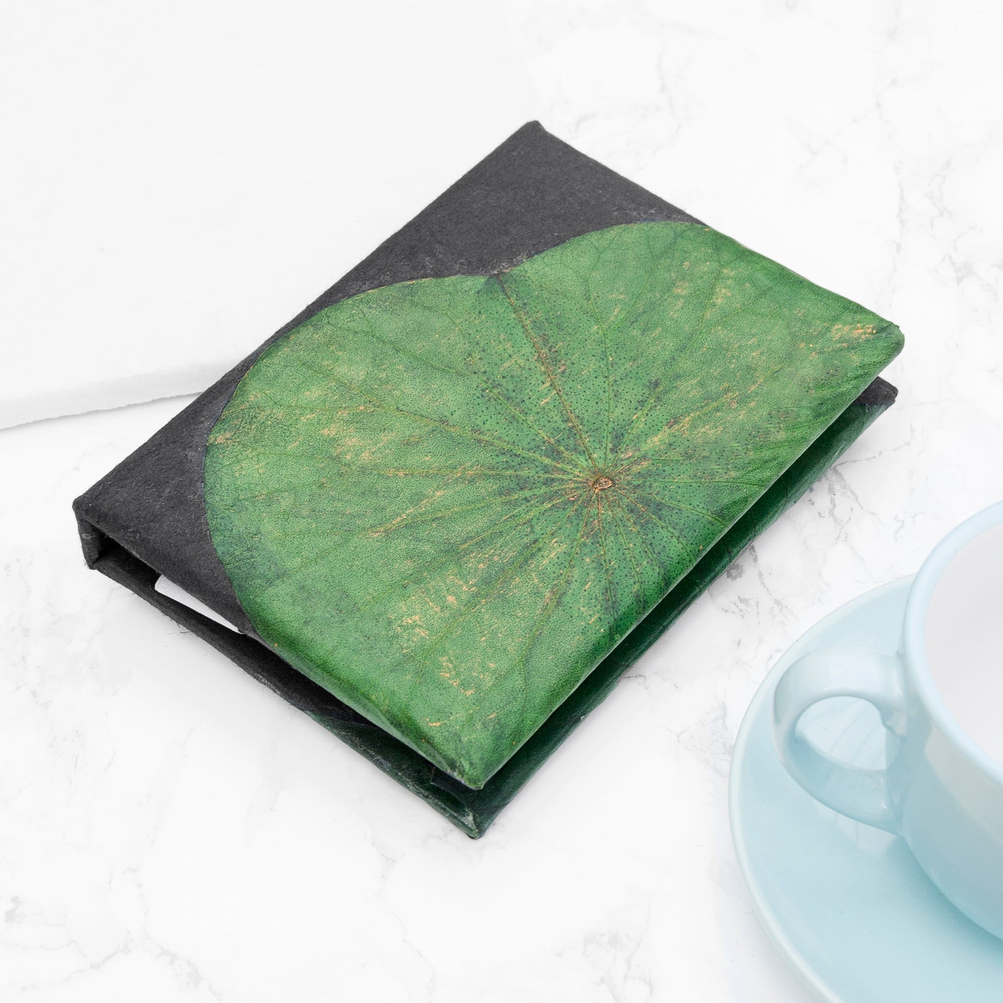Notebook with green leaf design on cover next to a cup and saucer on a white surface