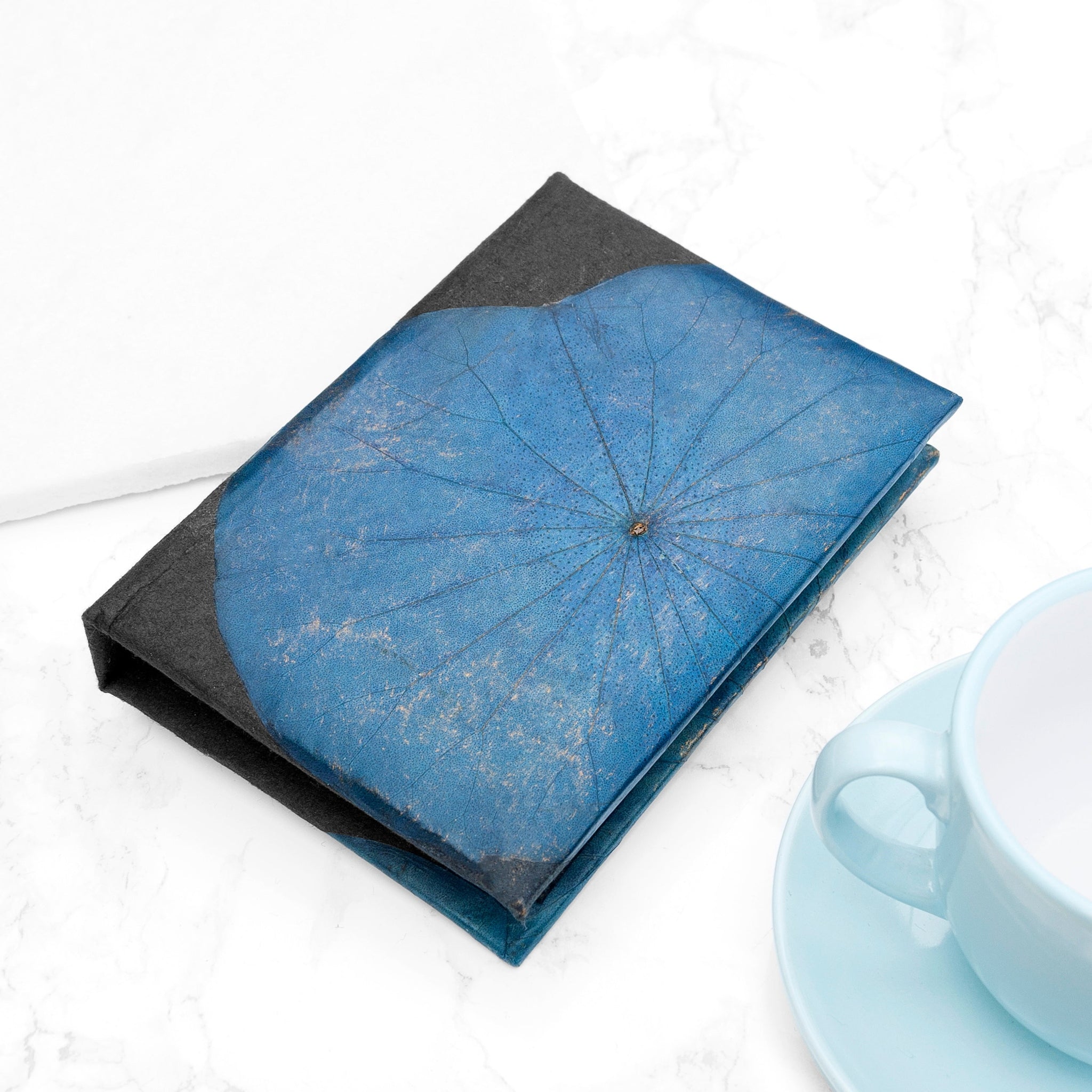 Blue dried flower on a black book with a light blue cup and saucer in the background
