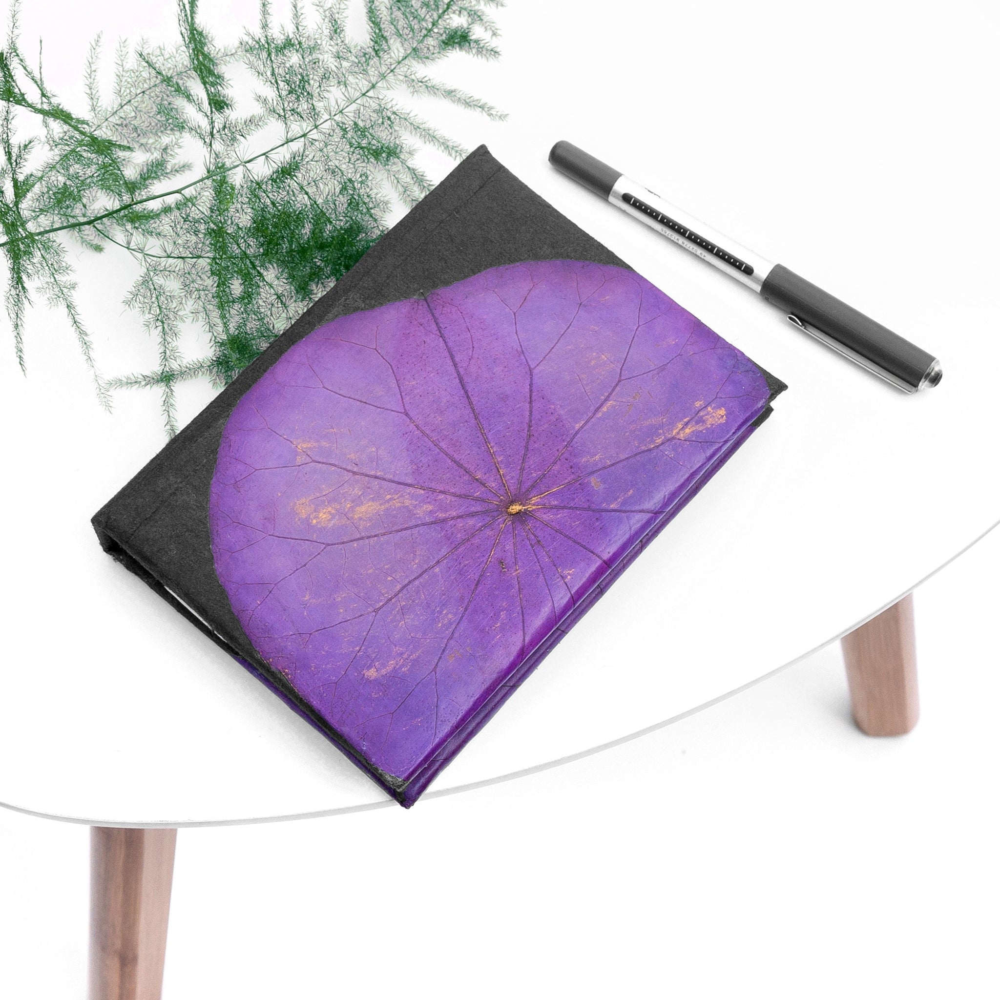 Purple leaf-patterned notebook with black cover on a white table, accompanied by a black pen.