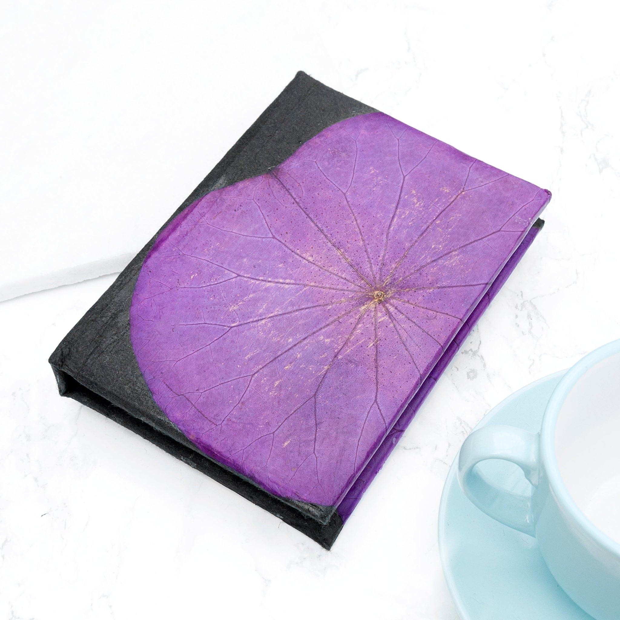 Purple notebook with black cover on a white surface next to a light blue mug.