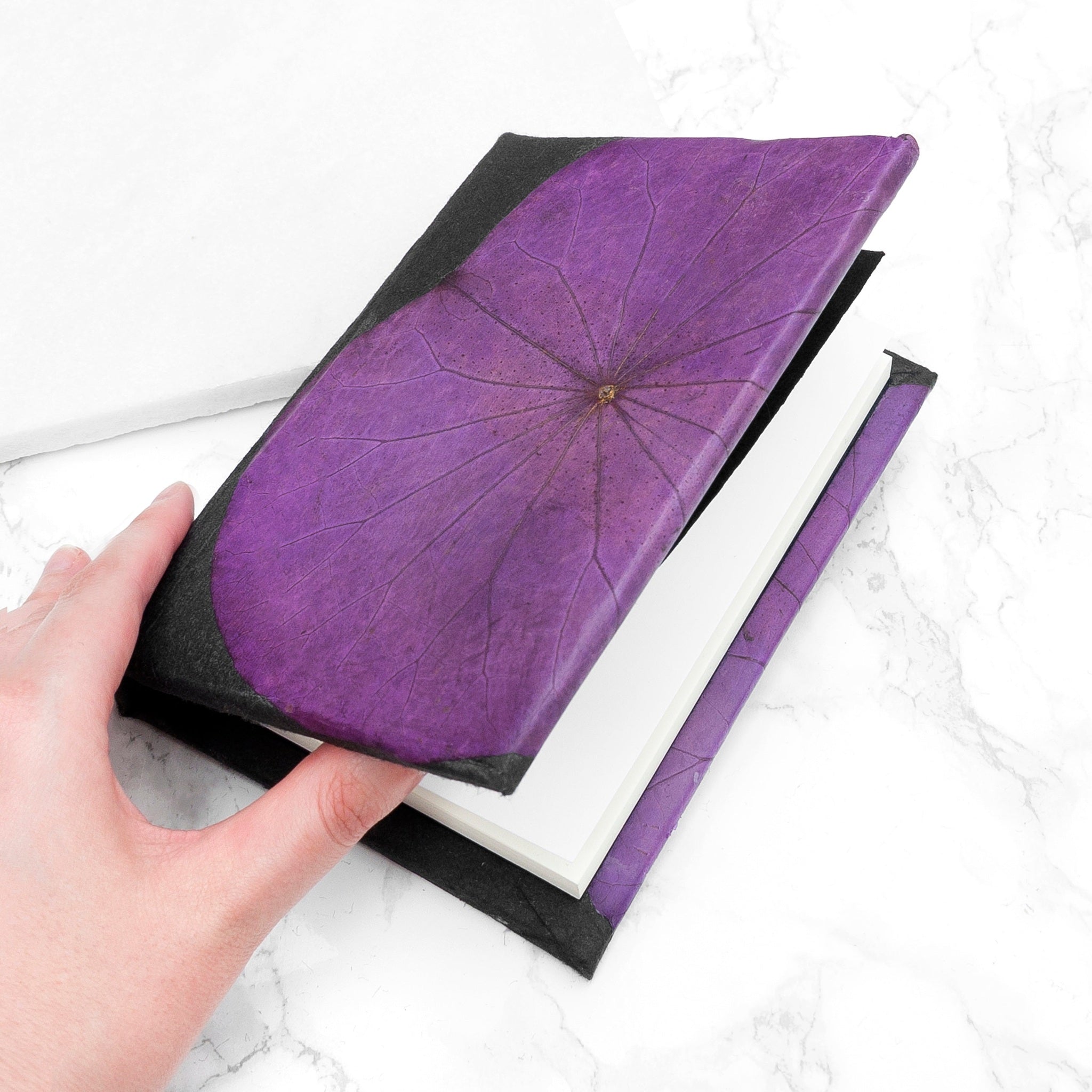 Hand holding a black book with a purple leaf cover on a white marble surface