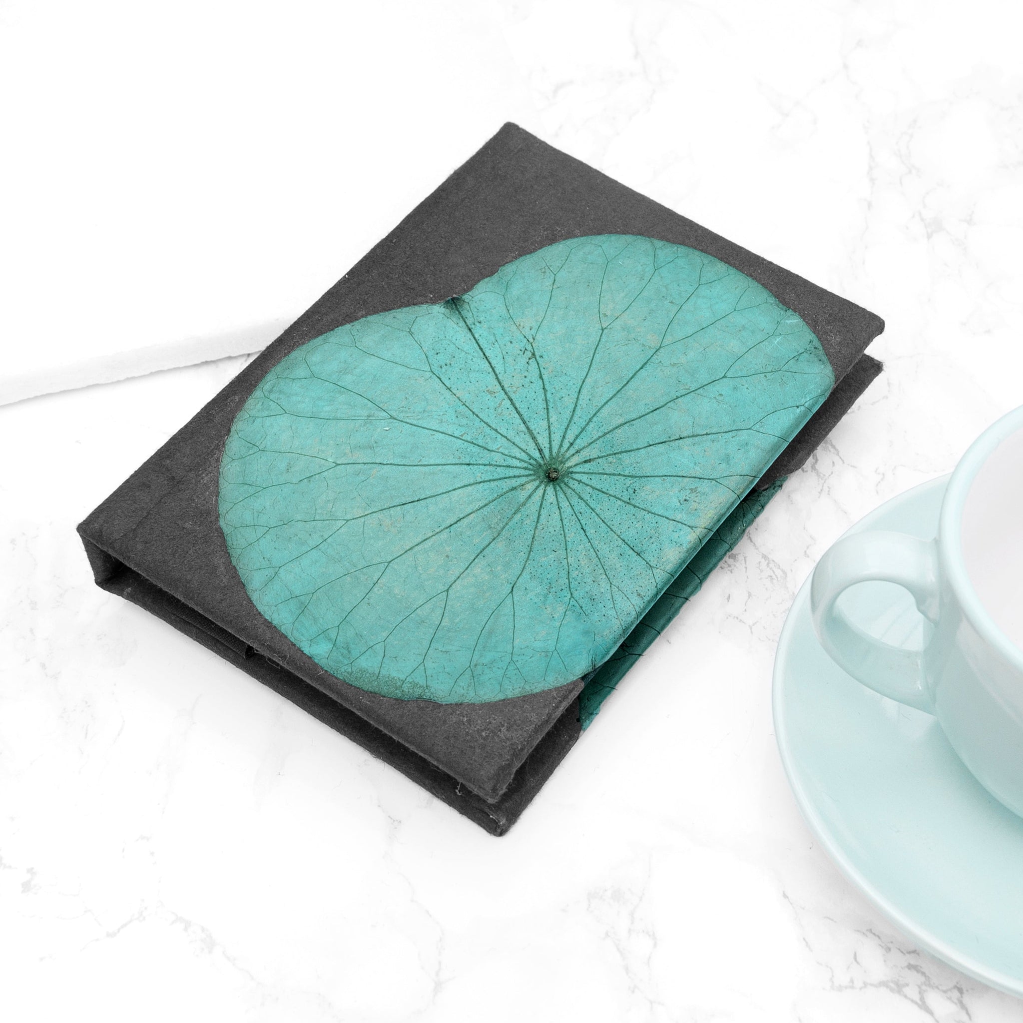 Teal flower design on a black notebook, placed on a white surface with a light blue cup and saucer.