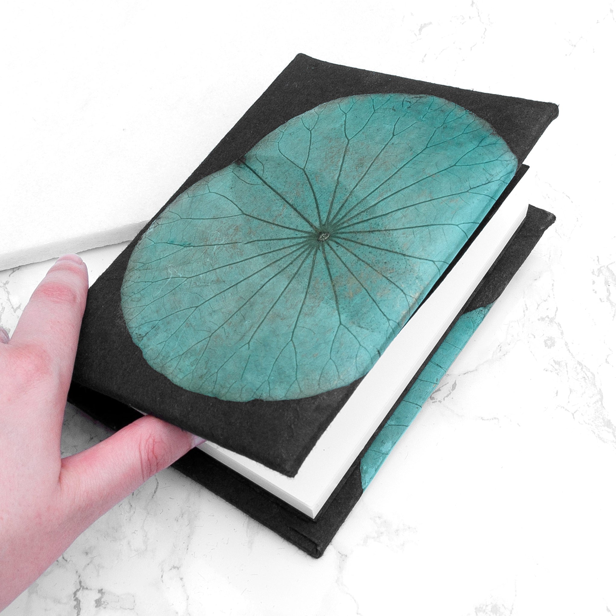 Hand holding a black book with a turquoise leaf design on a white marble surface