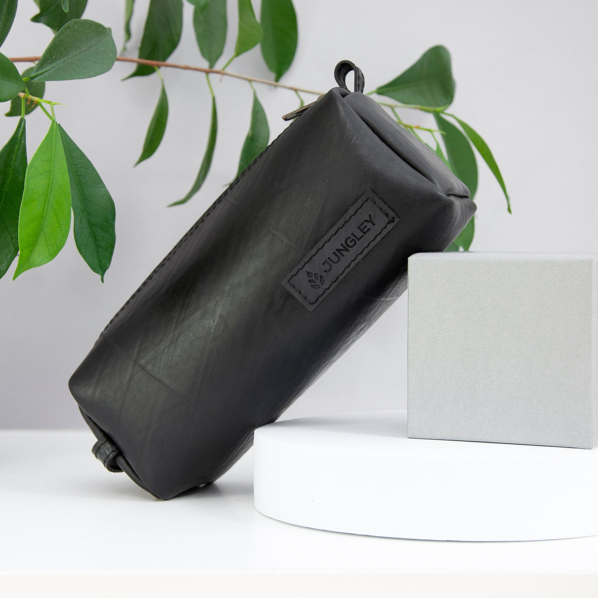 Black travel pouch with a brand logo on a white surface with green leaves in the background
