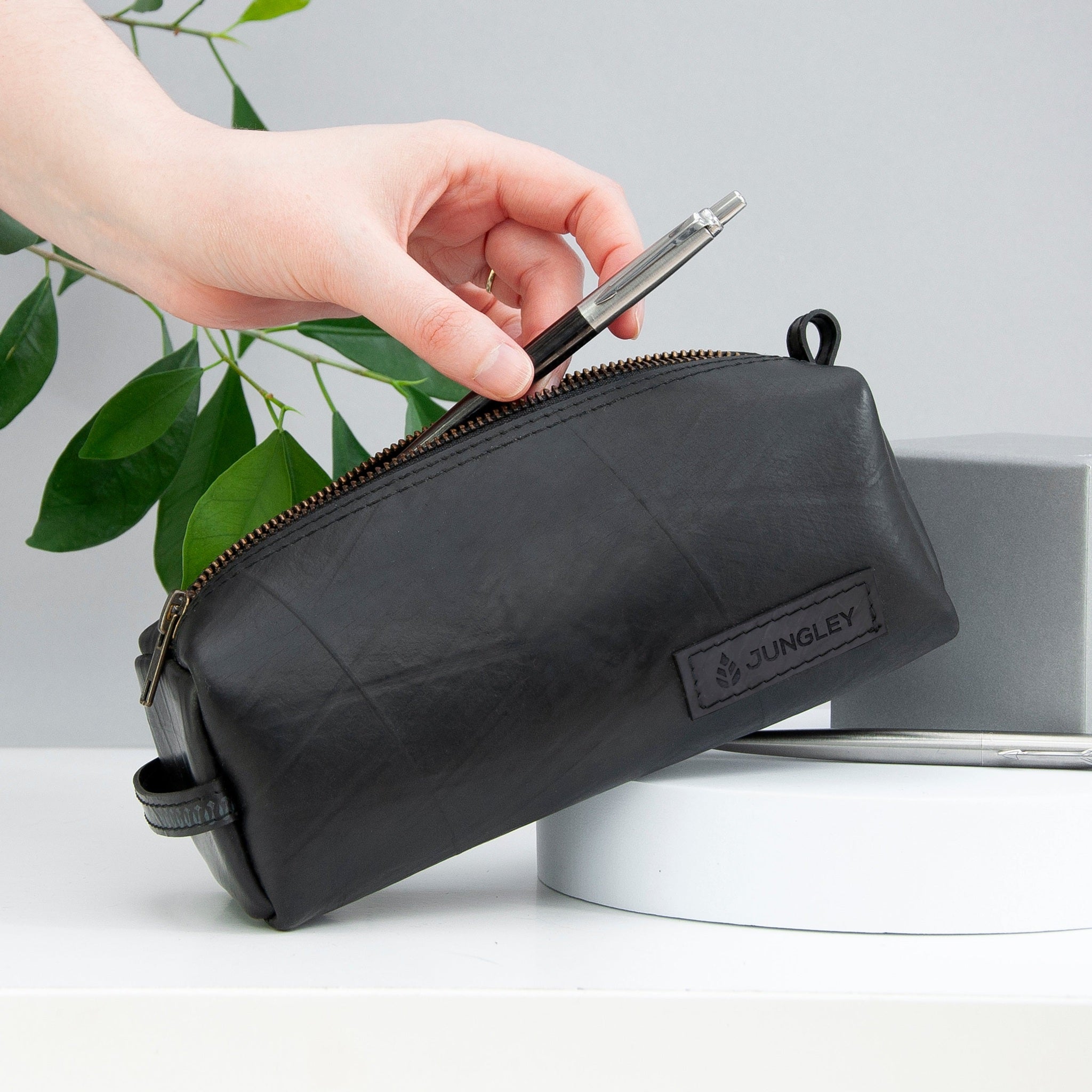 Person opening a black recycled rubber pencil case with 'Jungley' branding, holding a phone.