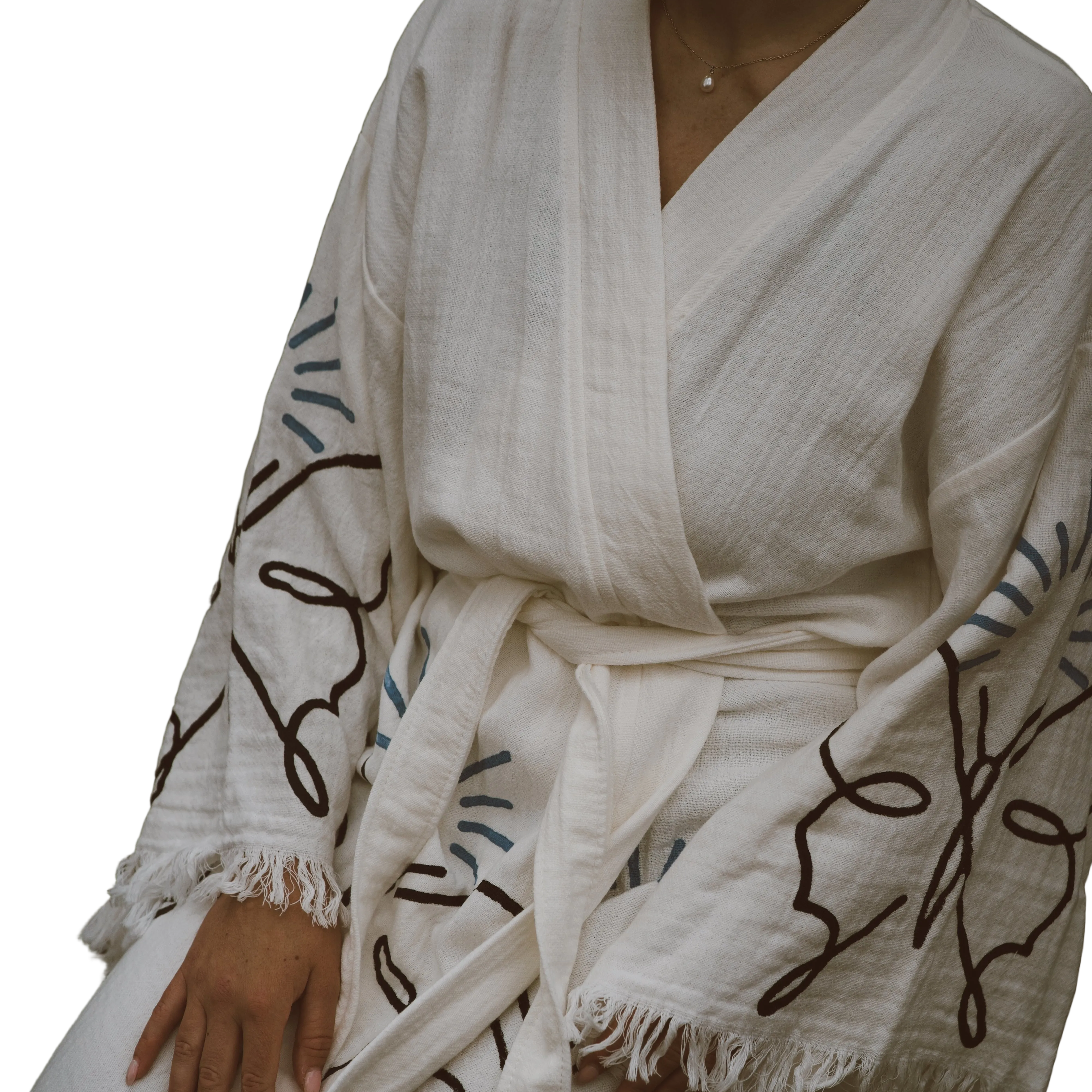 A close-up of a person wearing a cream-colored cotton robe with dark brown and blue abstract line patterns and fringed edges, shown against a plain white background.