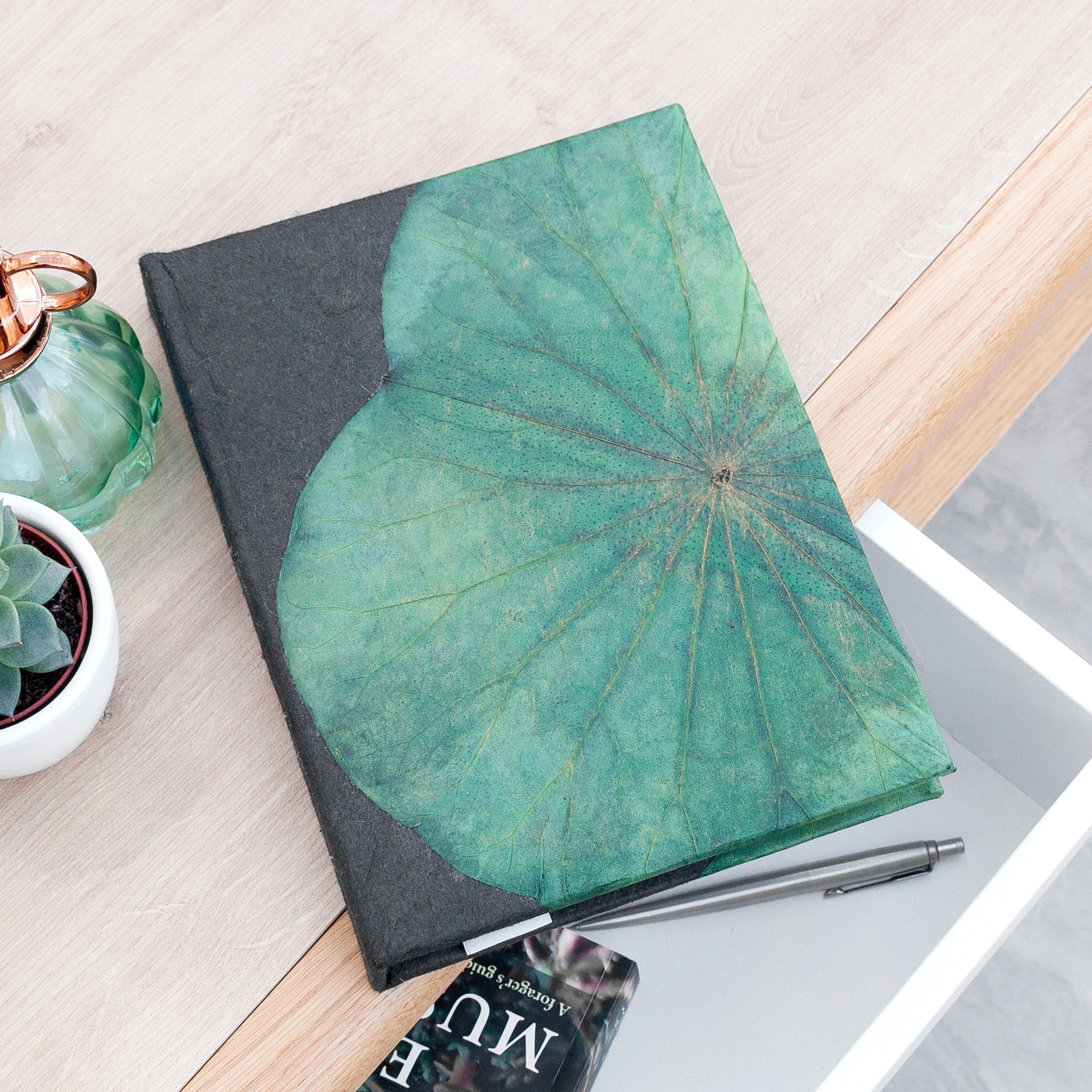 Notebook with a green leaf design on a desk with a pen and plant