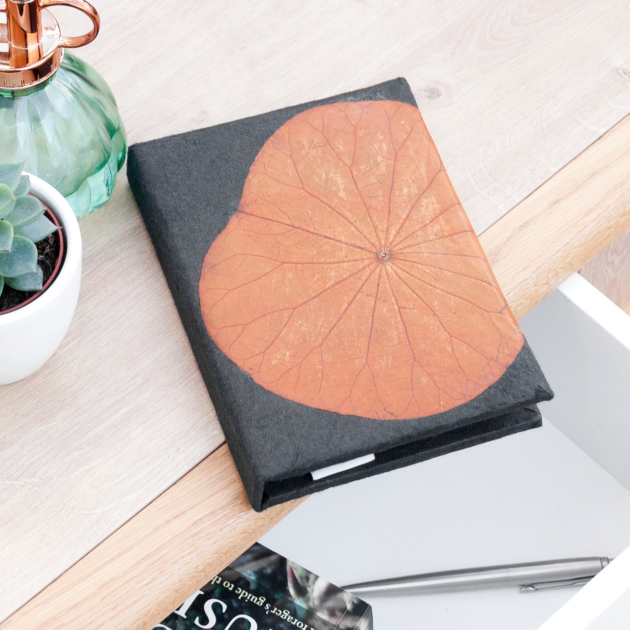 Notebook with a brown leaf design on a white surface with a plant and magazine.