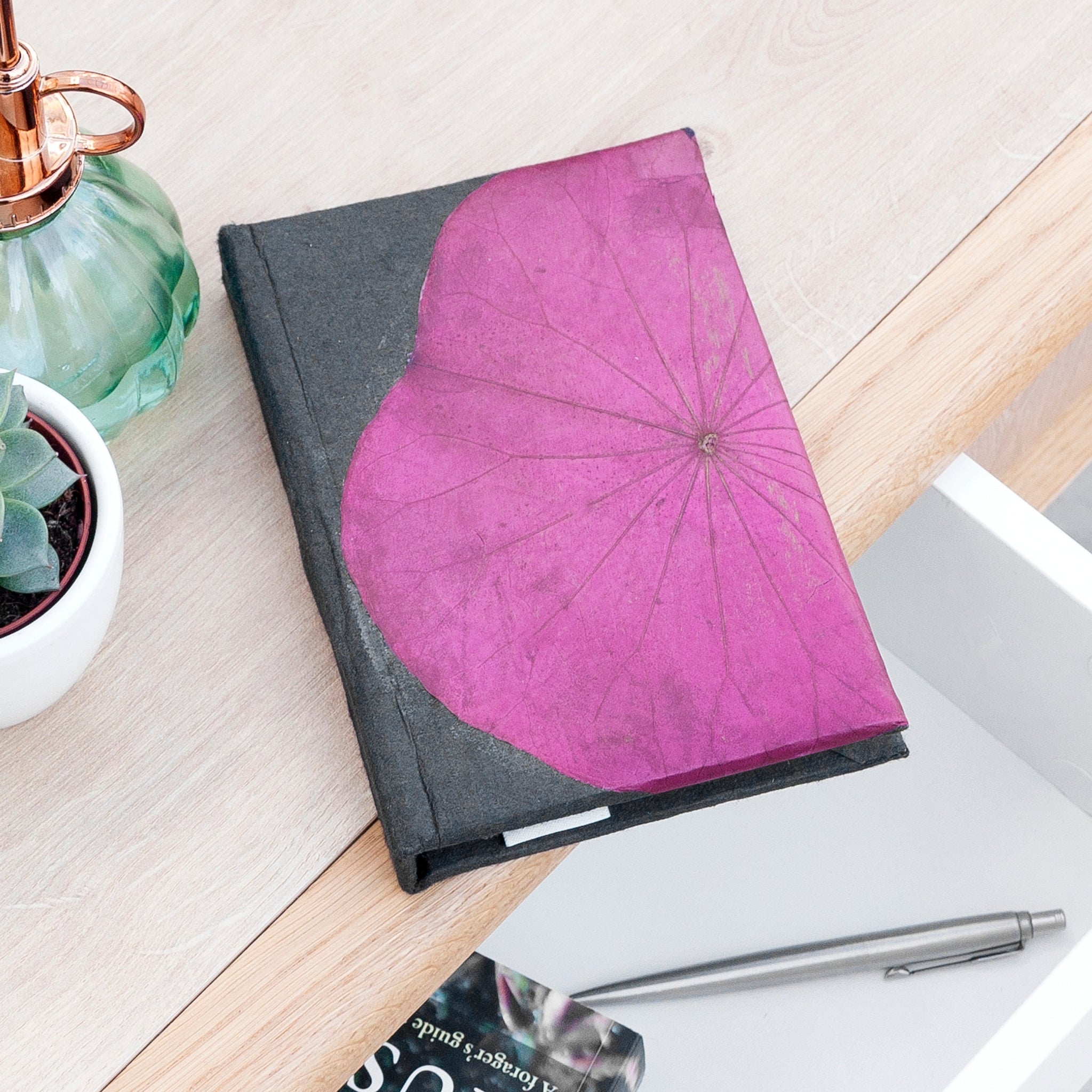Notebook with a pink heart-shaped cover on a desk with a pen and plant