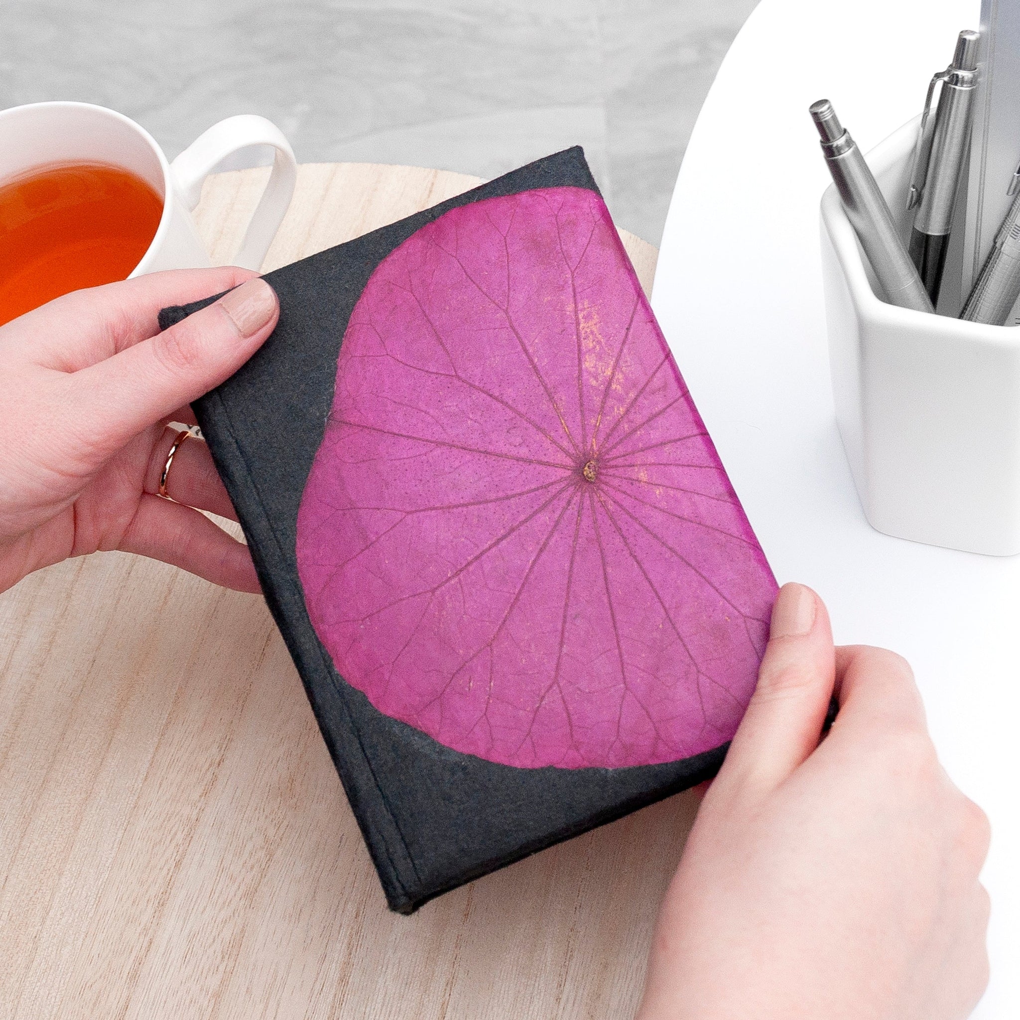 Person holding a notebook with a purple leaf design on a desk with a cup of tea and pens.