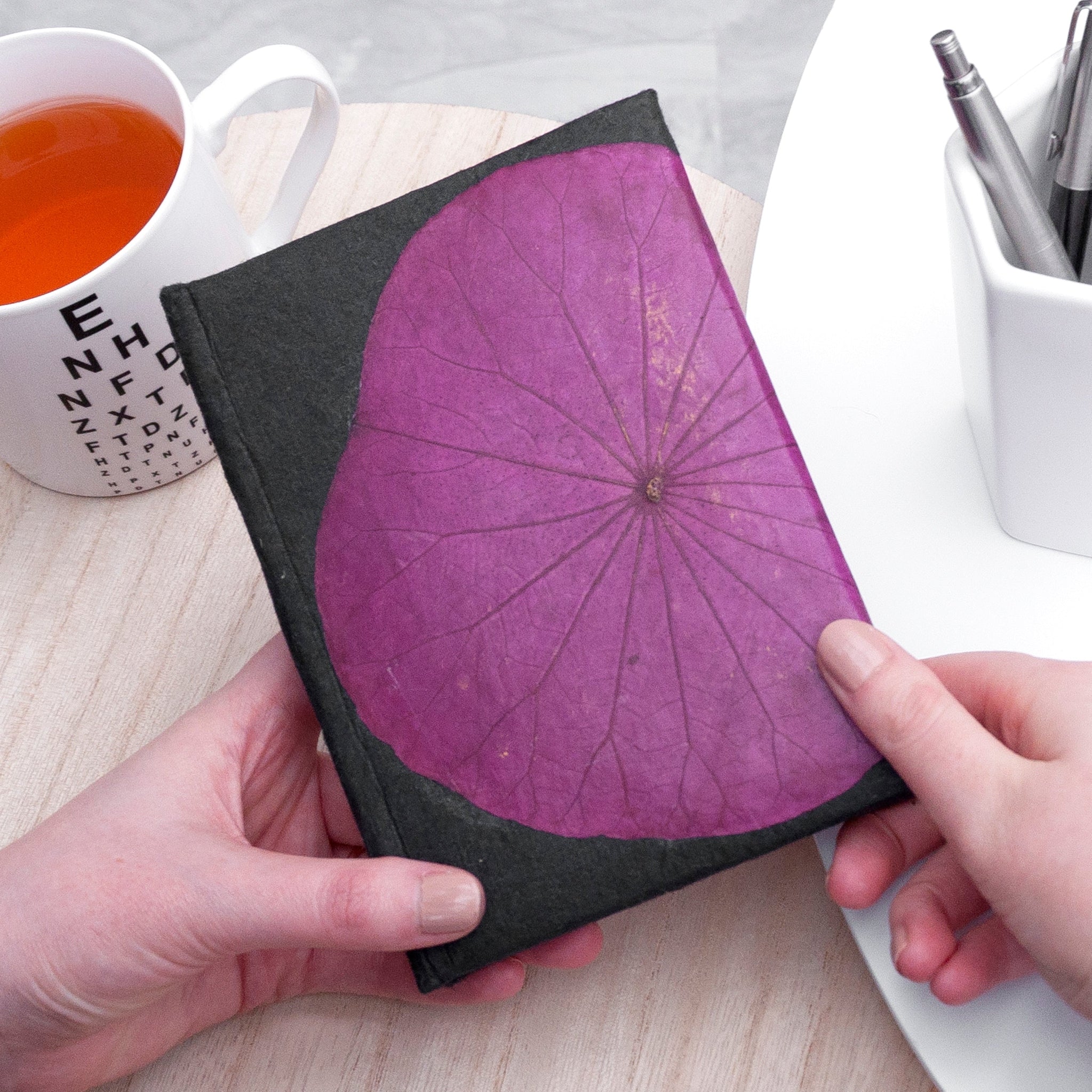 Person holding a notebook with a purple leaf design on a table with a cup of tea and stationery.