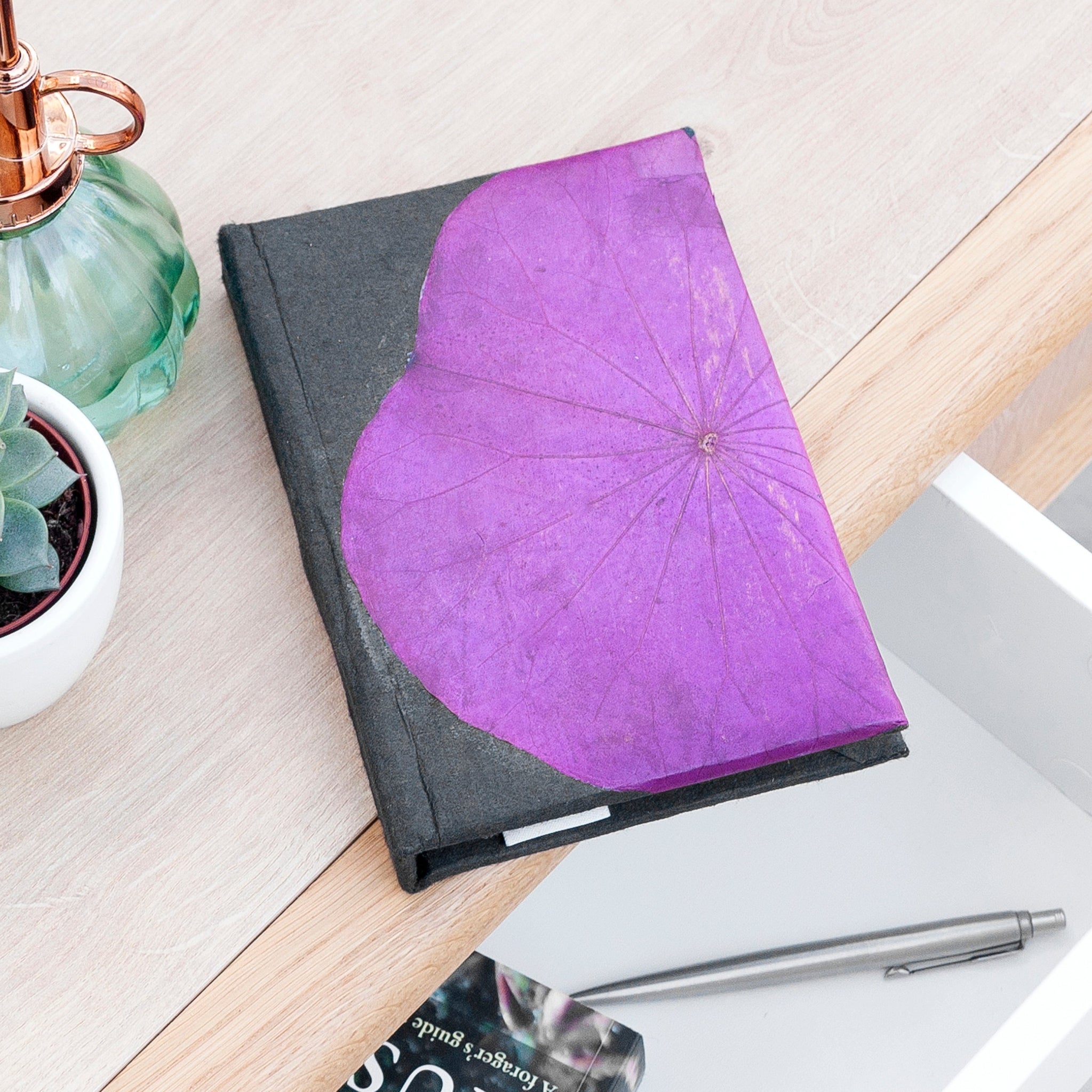 Notebook with a purple heart-shaped cover on a desk with a pen and plant