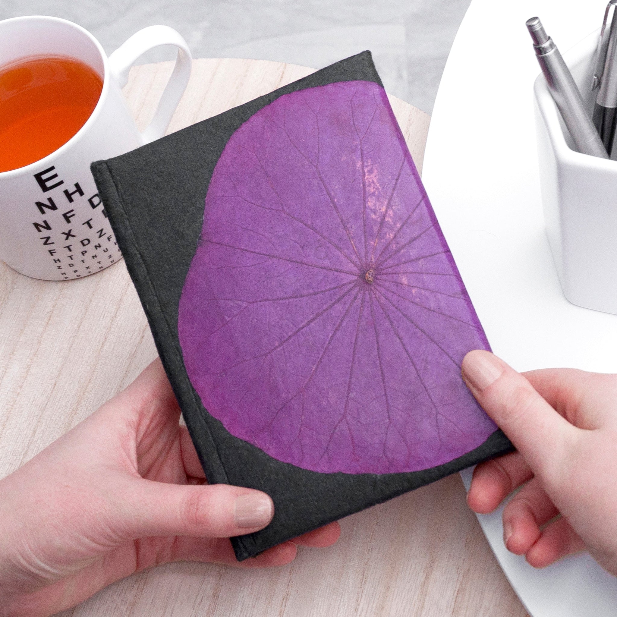 Notebook with purple leaf design held by hands on a desk with a cup of tea and stationery.
