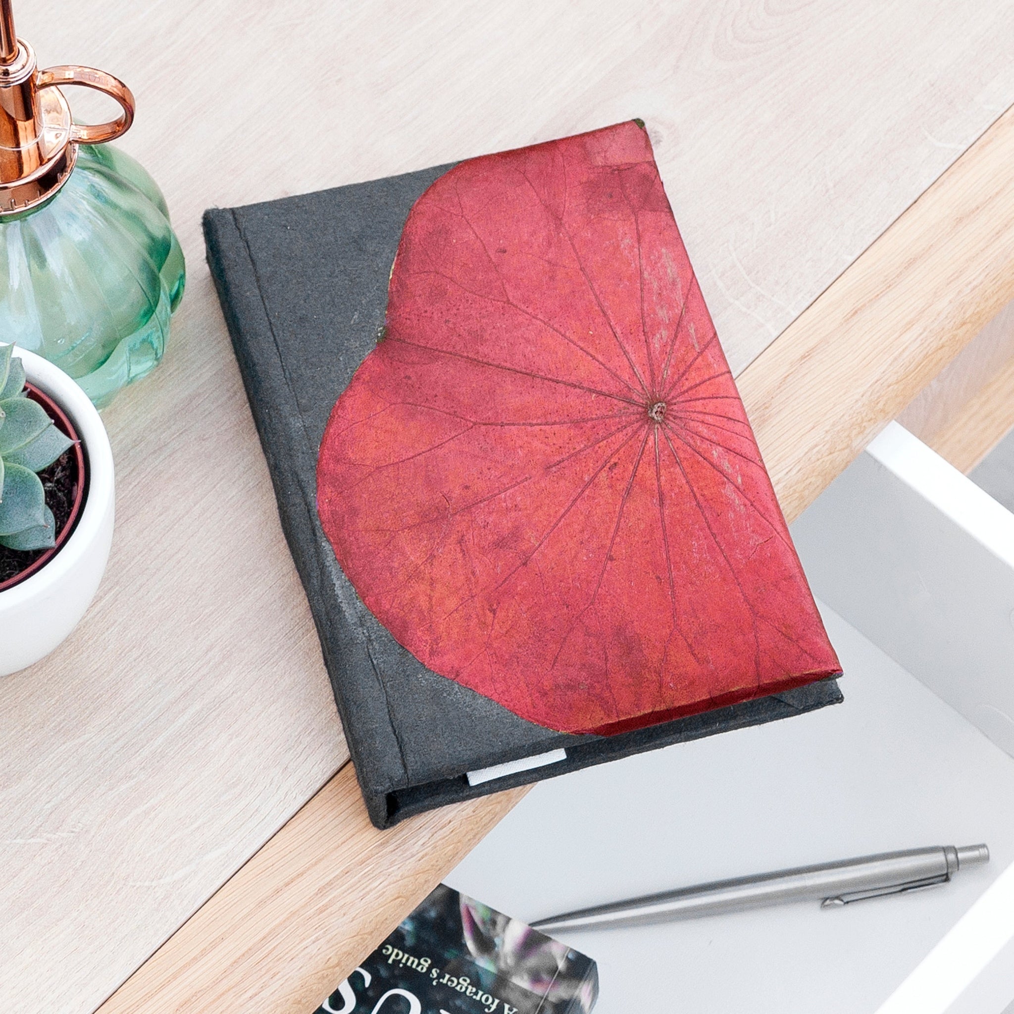 Notebook with red leaf design on cover, placed on a wooden surface with a pen and plant in the background.