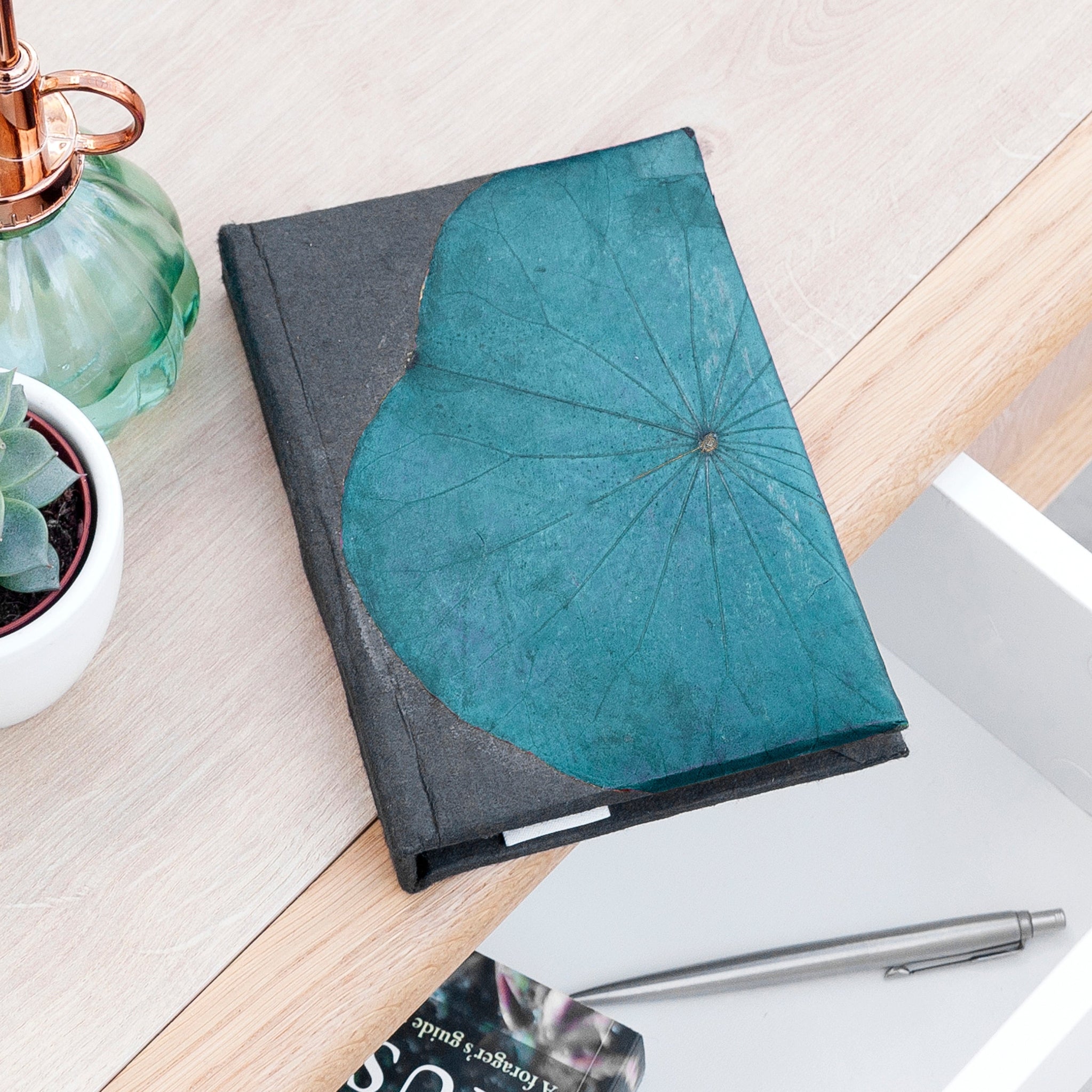 Notebook with teal leaf design on a wooden surface with a pen and plant.