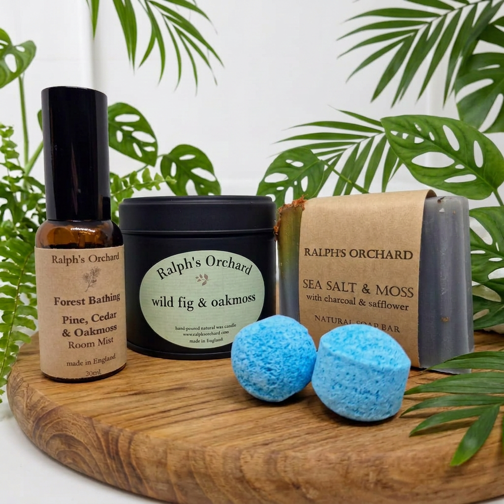 A spa gift set from Ralph's Orchard arranged on a round wooden tray with tropical green leaves in the background. The set includes a forest bathing room mist bottle, a wild fig & oakmoss tin candle, a sea salt & moss natural soap bar, and two blue bath bombs.