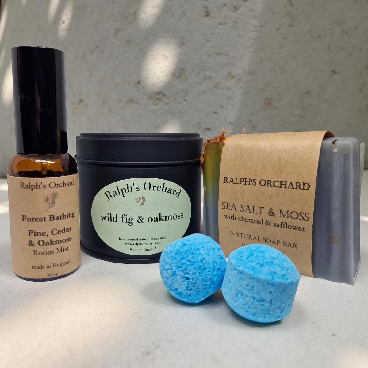 Collection of Ralph's Orchard products including a bottle, jar, soap bar, and bath bombs on a textured surface.