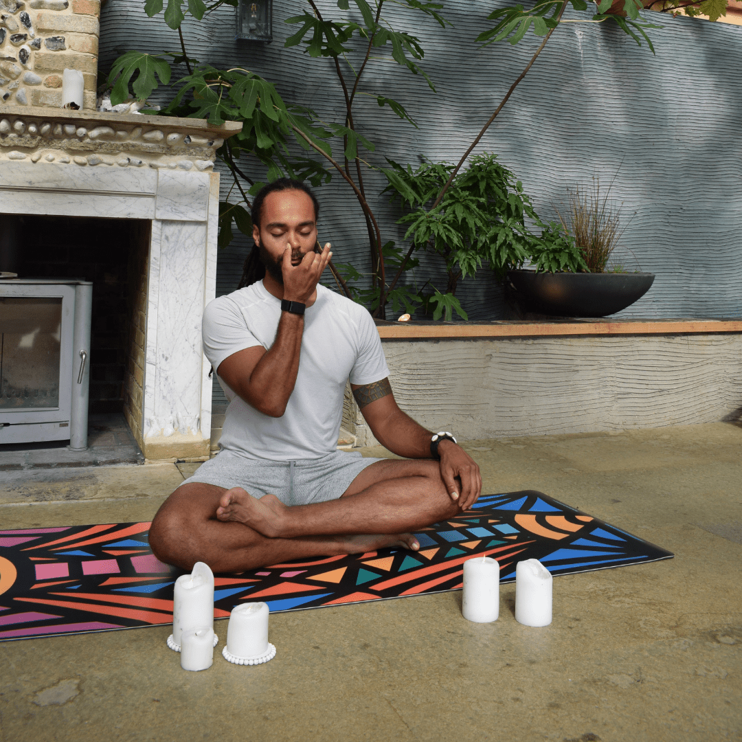 Man sitting in a meditative pose on a colorful mat with candles around him, surrounded by plants and a fireplace.