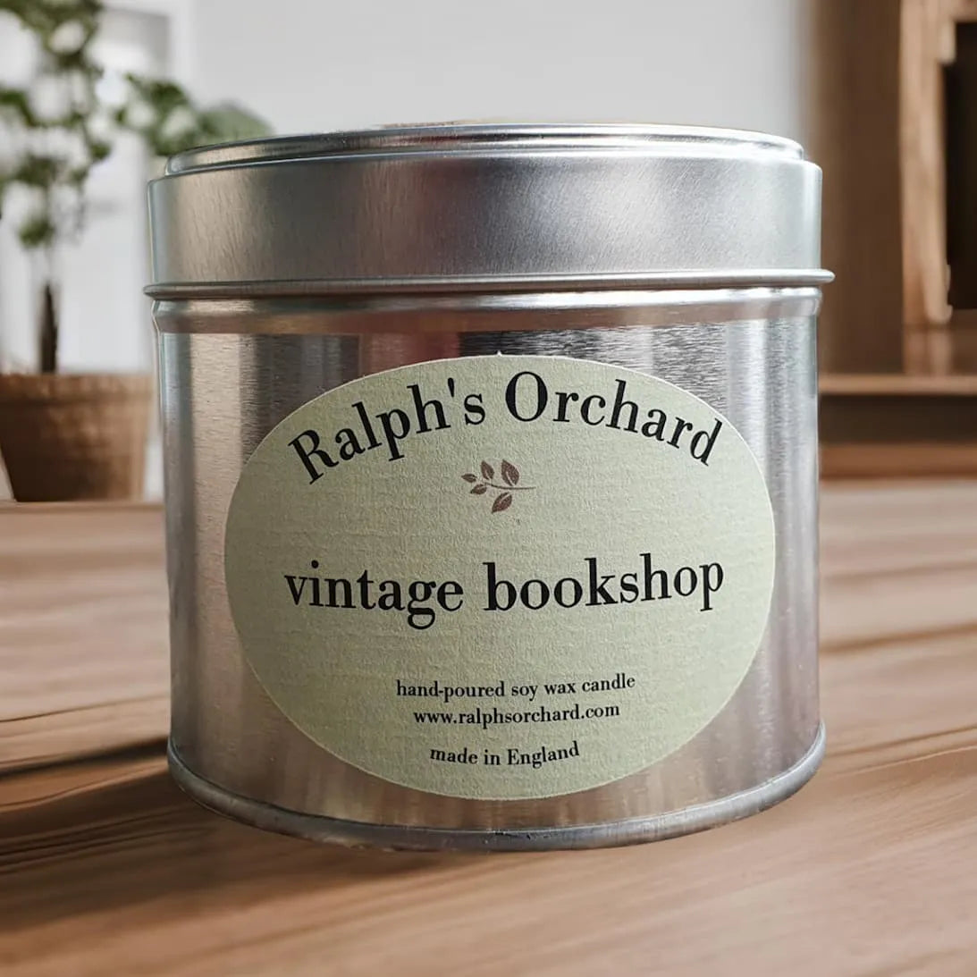 Candle in a metal tin with 'Ralph's Orchard vintage bookshop' label on a wooden surface.
