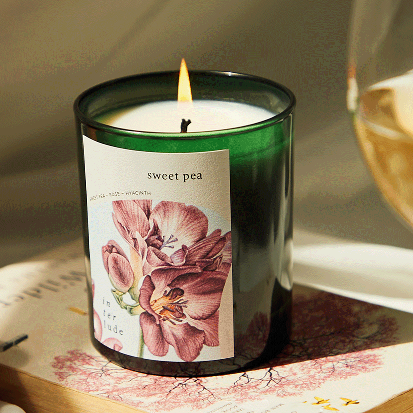 A green glass candle jar with a lit flame, featuring a label that reads 'sweet pea' and shows pink floral artwork. The candle is styled on a book next to a glass of white wine.