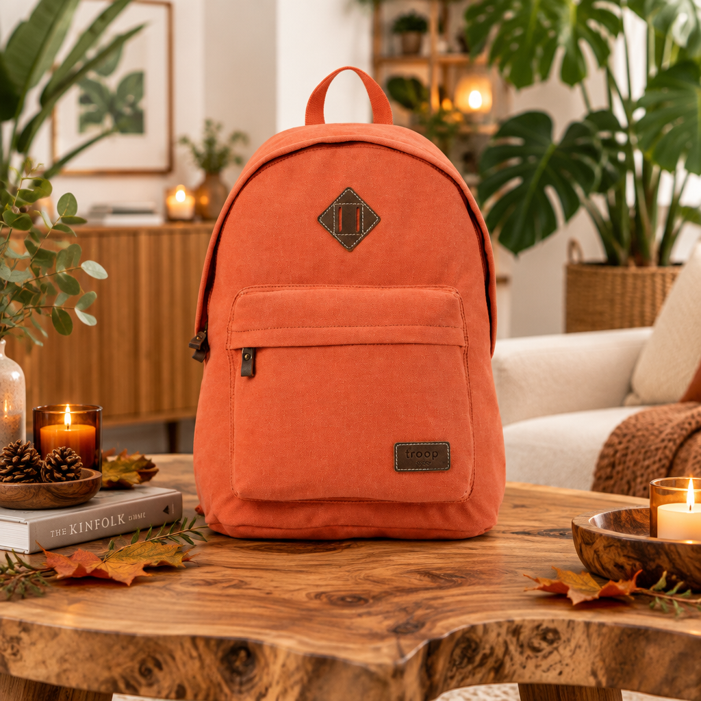 An orange canvas backpack with dark brown leather accents sits on a rustic wooden table in a cosy, warmly lit living room setting with plants and candles.