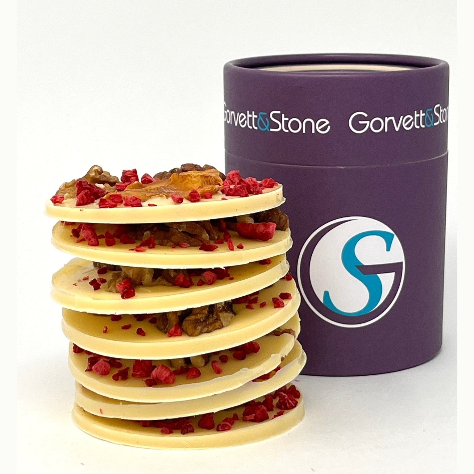 Stack of white chocolate discs with red fruits next to a purple Gorrett & Stone container on a white background