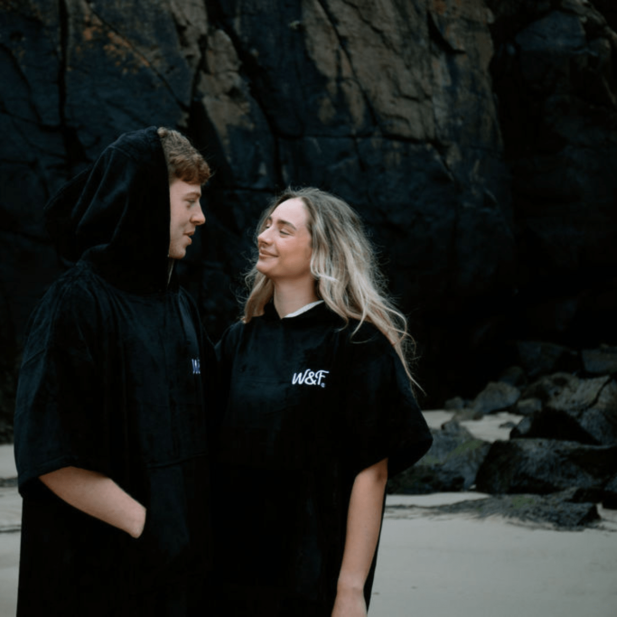 Two people standing on a rocky beach, one wearing a black towel robe and the other with 'WST' logo.