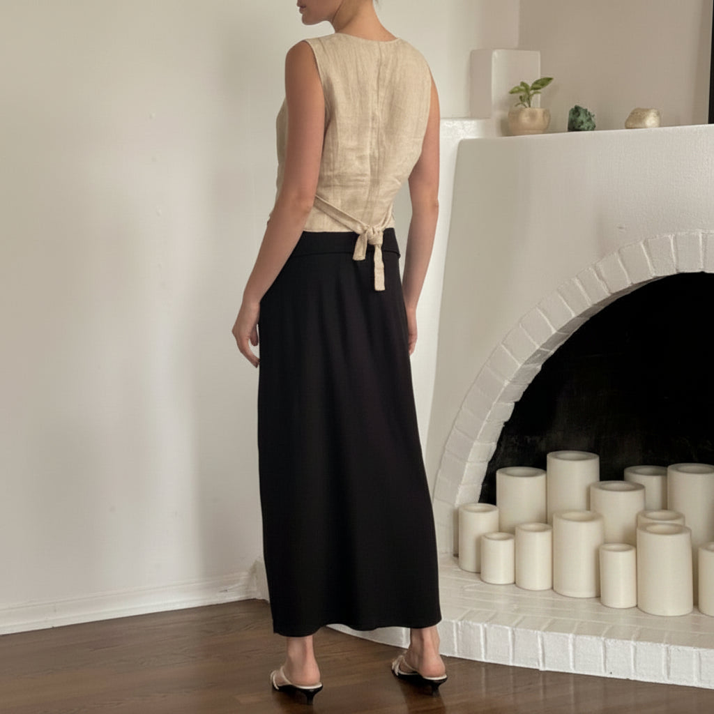 Person wearing a beige sleeveless top and black skirt in a room with white walls and decorative items.