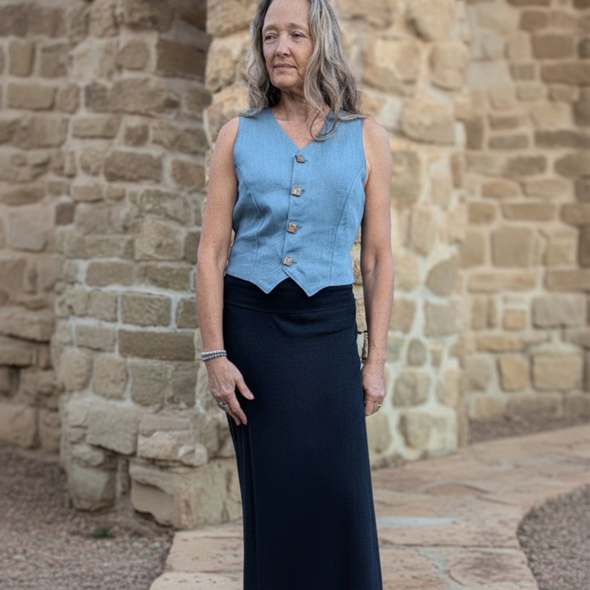 Woman wearing a blue vest over a black dress against a stone wall.