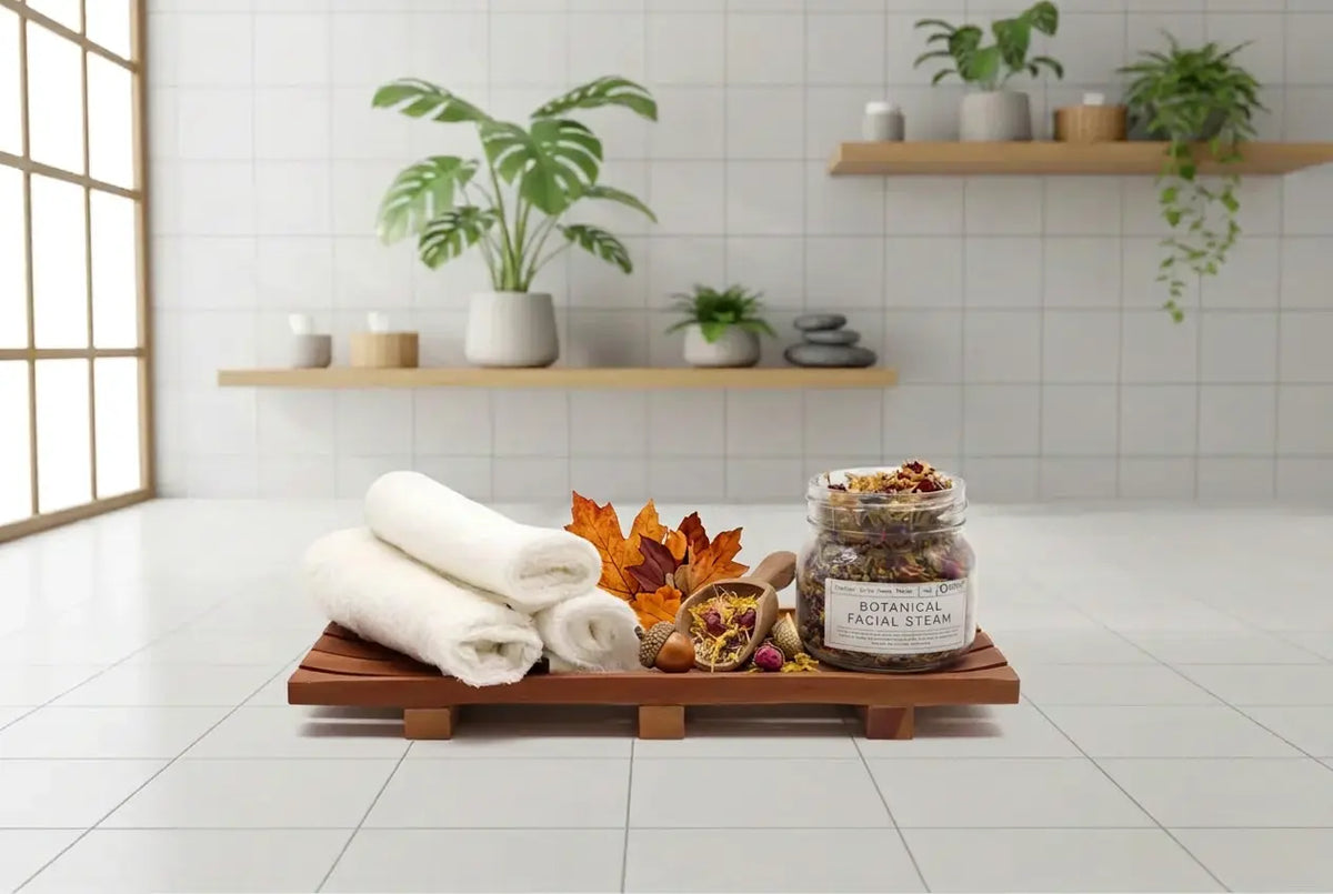 A wooden spa tray holding three rolled white towels, dried orange autumn leaves, and a glass jar labeled 'Botanical Magic Steam' sits on a white tiled floor in a bright, modern bathroom or spa room with shelves and plants in the background.