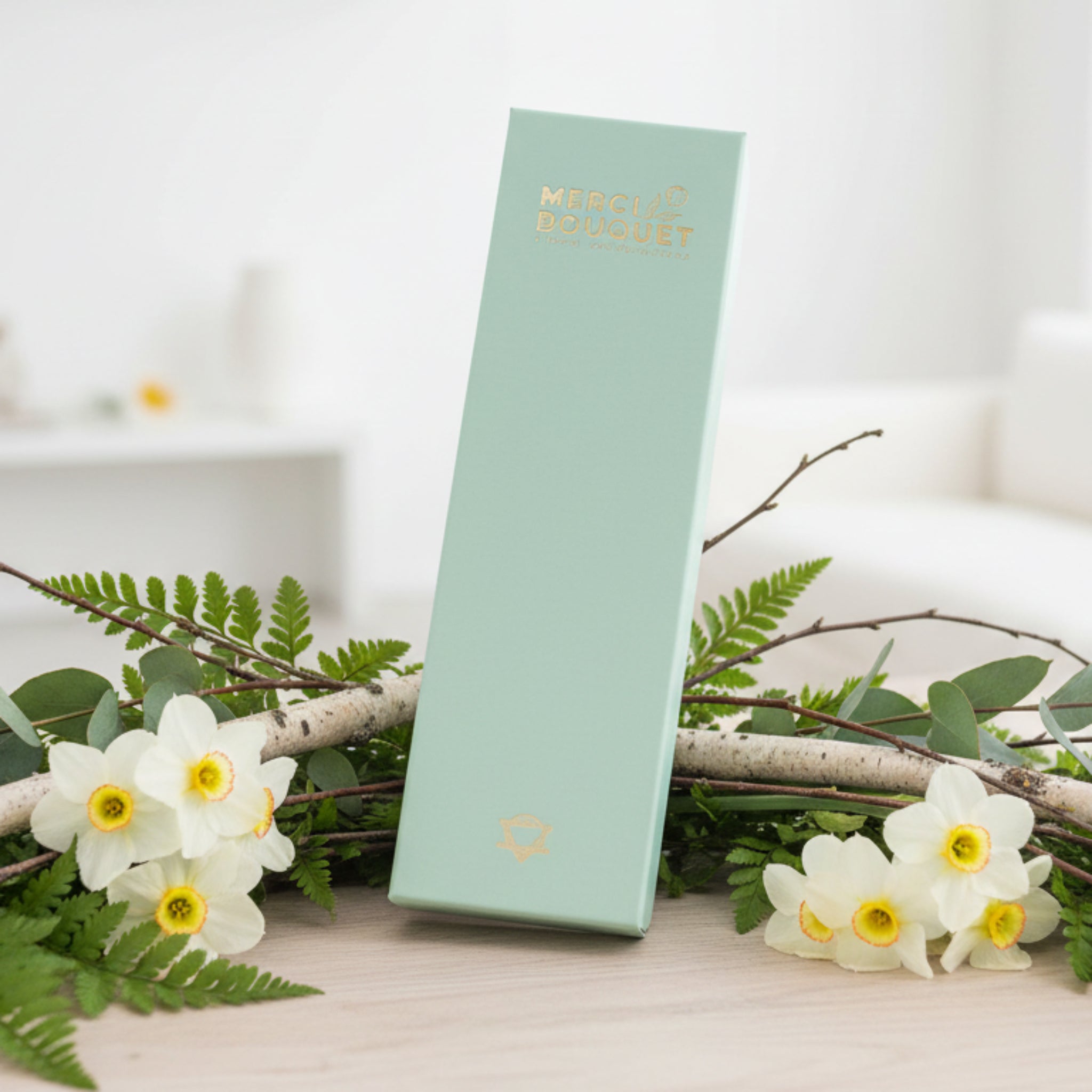 Mint green product box with gold text on a white surface with greenery and flowers.