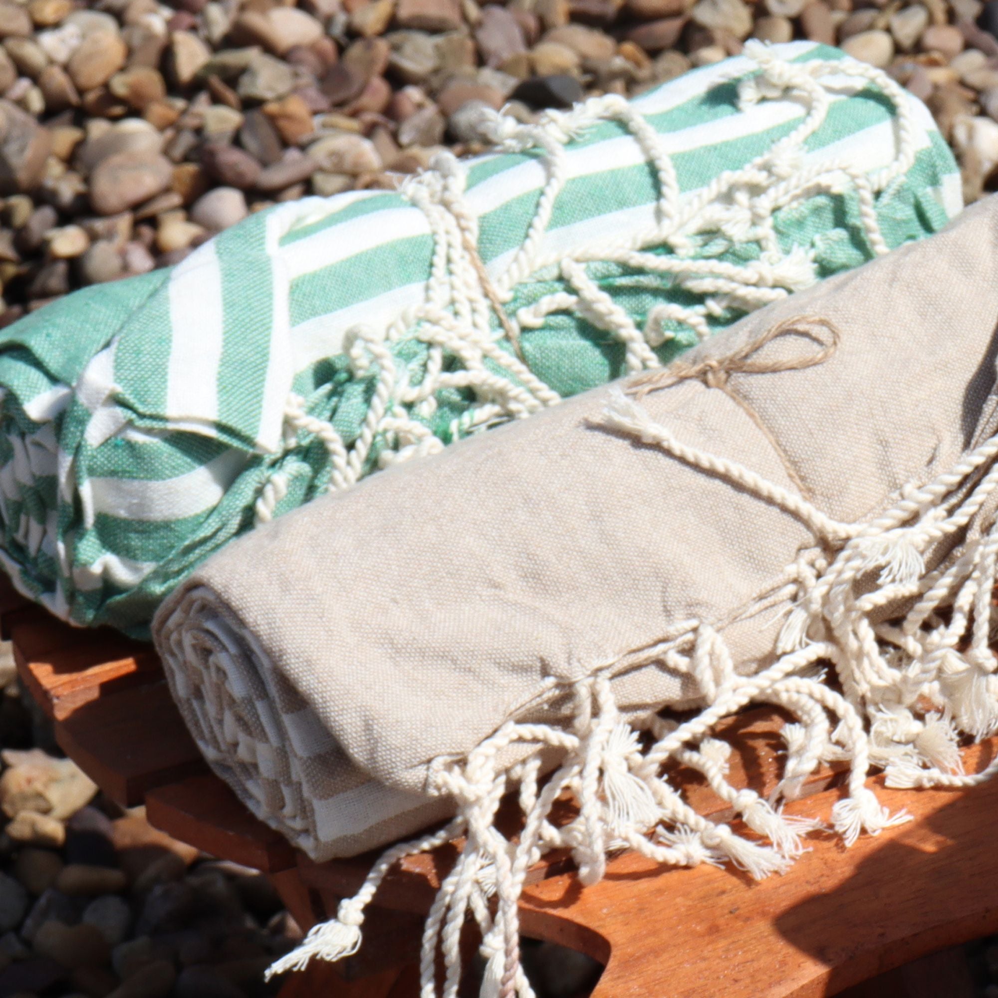 Two folded towels, one green and white patterned, the other beige with tassels, on a wooden stand with a stone background.