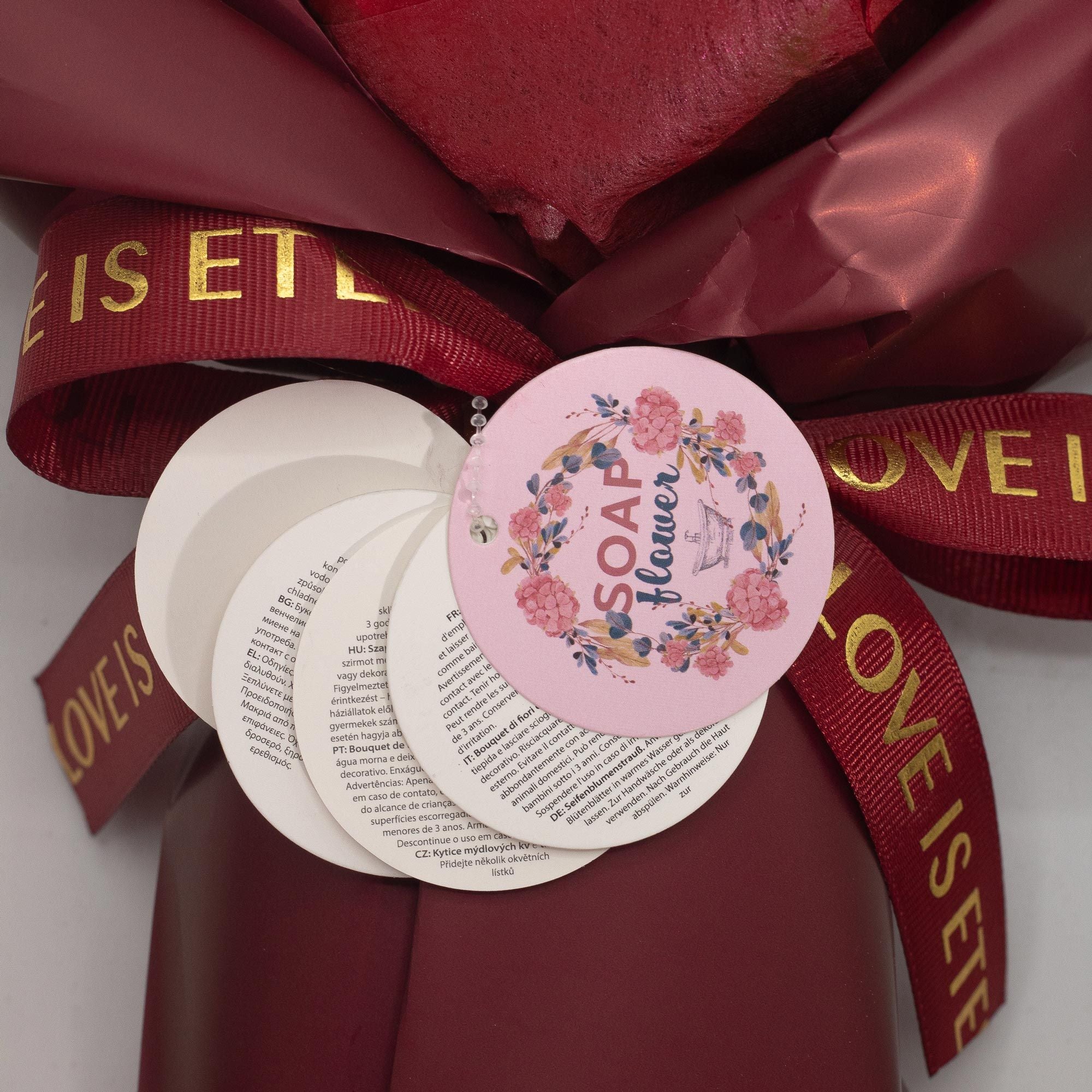 Pink soap with floral design and text, surrounded by red ribbons with gold text on a white background